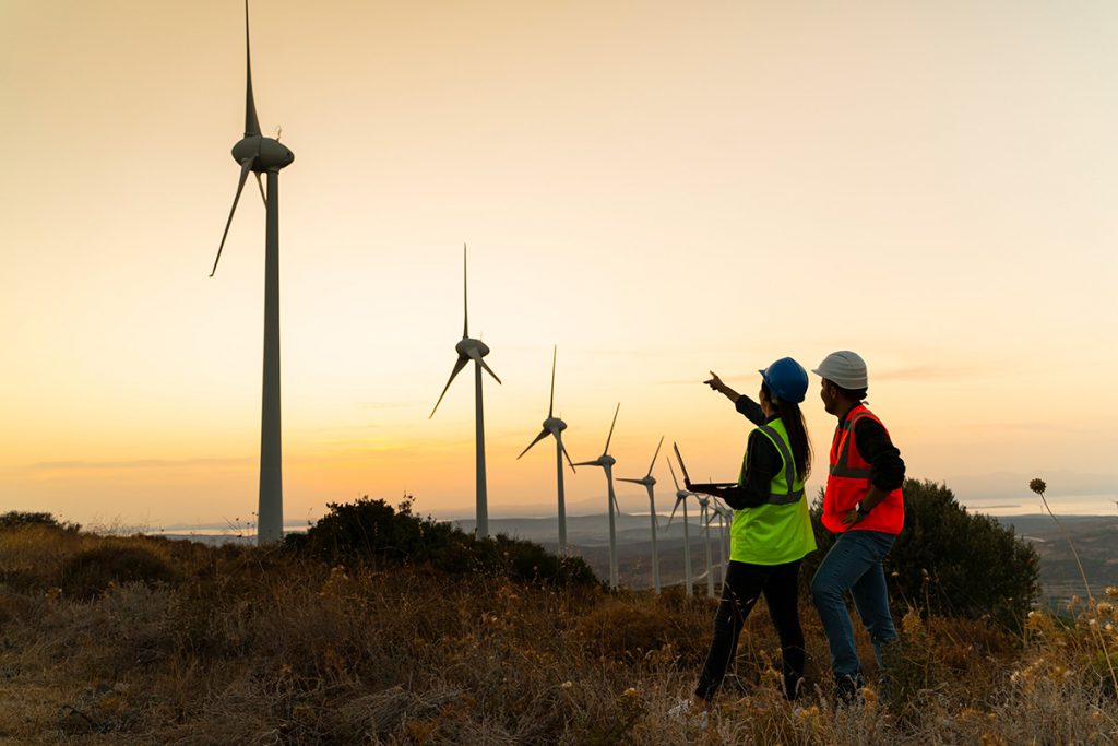 Silhouette of young engineer holding laptop computer planning and working for the energy industry and standing beside a wind turbines farm power station at sunset time